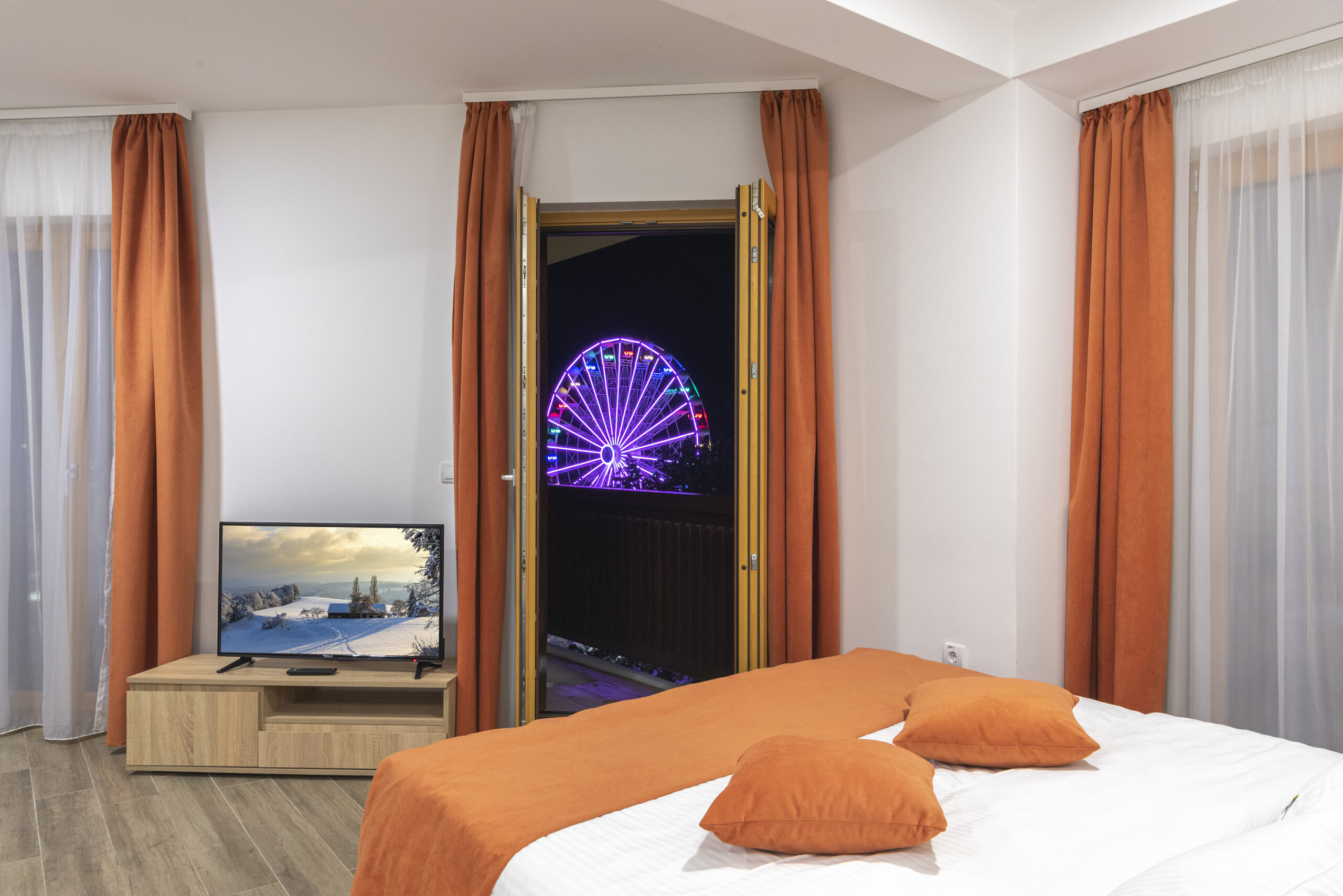 Komforna hotelska soba sa narandžastim detaljima i otvorenom terasom sa koje se pruža pogled na osvetljeni panoramski točak noću.